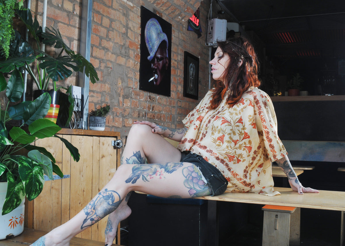 A woman with tattoos sits on a wooden counter in a cafe, wearing a cream-colored bohemian patterned blouse and black denim shorts.