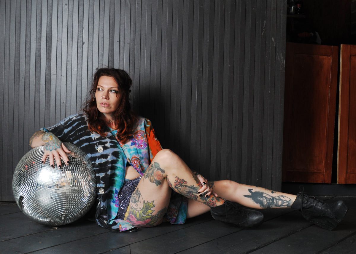 A tattooed woman sits on a dark wooden floor against a dark paneled wall, holding a large silver disco ball. She is wearing a colorful tie-dye shirt and black boots.