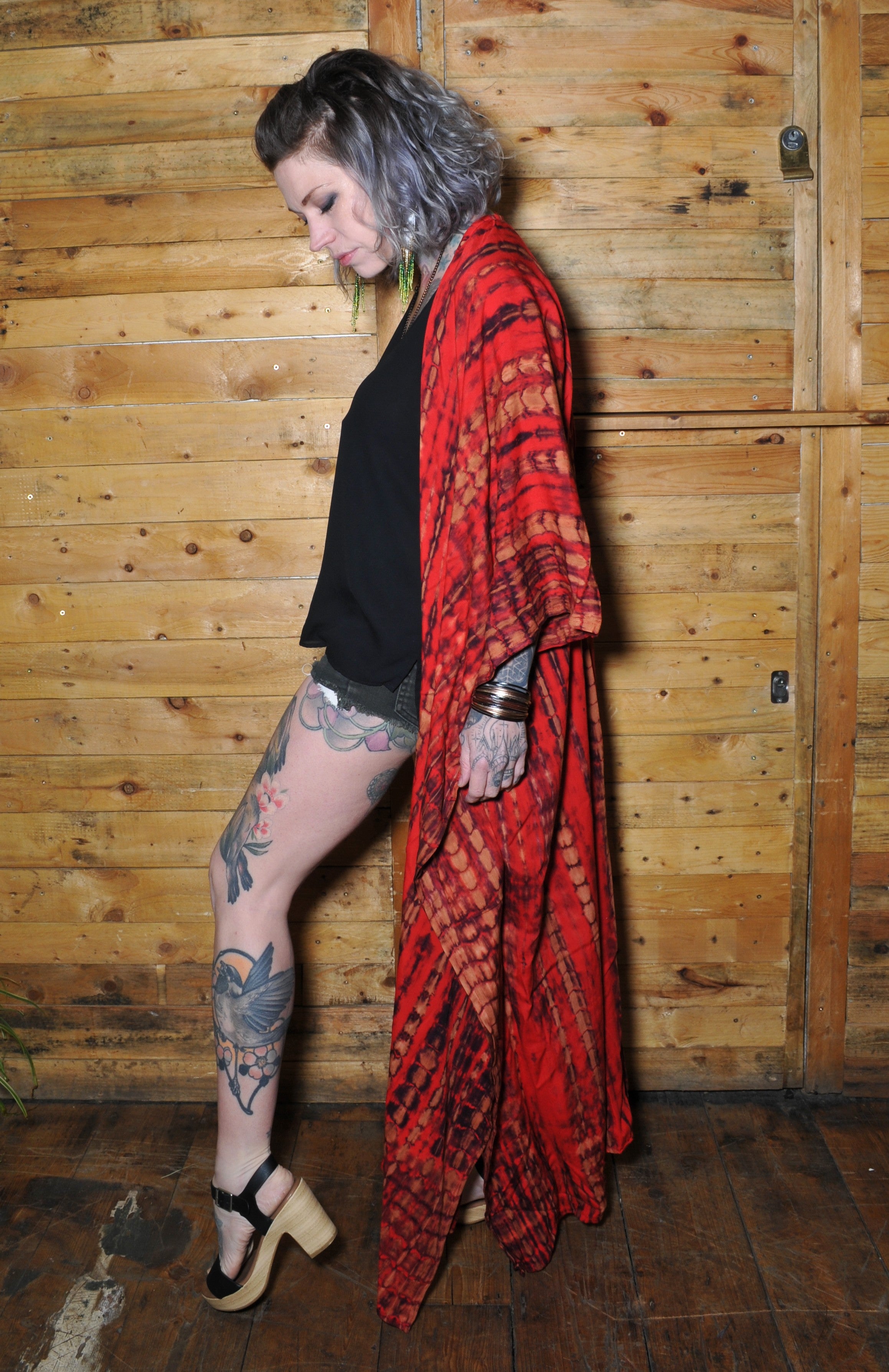 Model wearing a red and black tie dyed kimono standing up against a wooden retro bar wearing wooden platform heels 